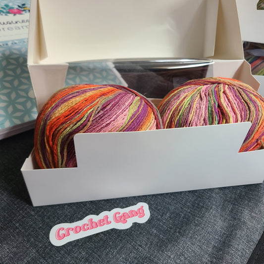 Two large balls of variegated crochet yarn in shades of orange, pink, purple, green, and yellow, displayed inside an open white cardboard box. A sticker reading 'Crochet Gang' is visible in the foreground on a dark gray textured surface.