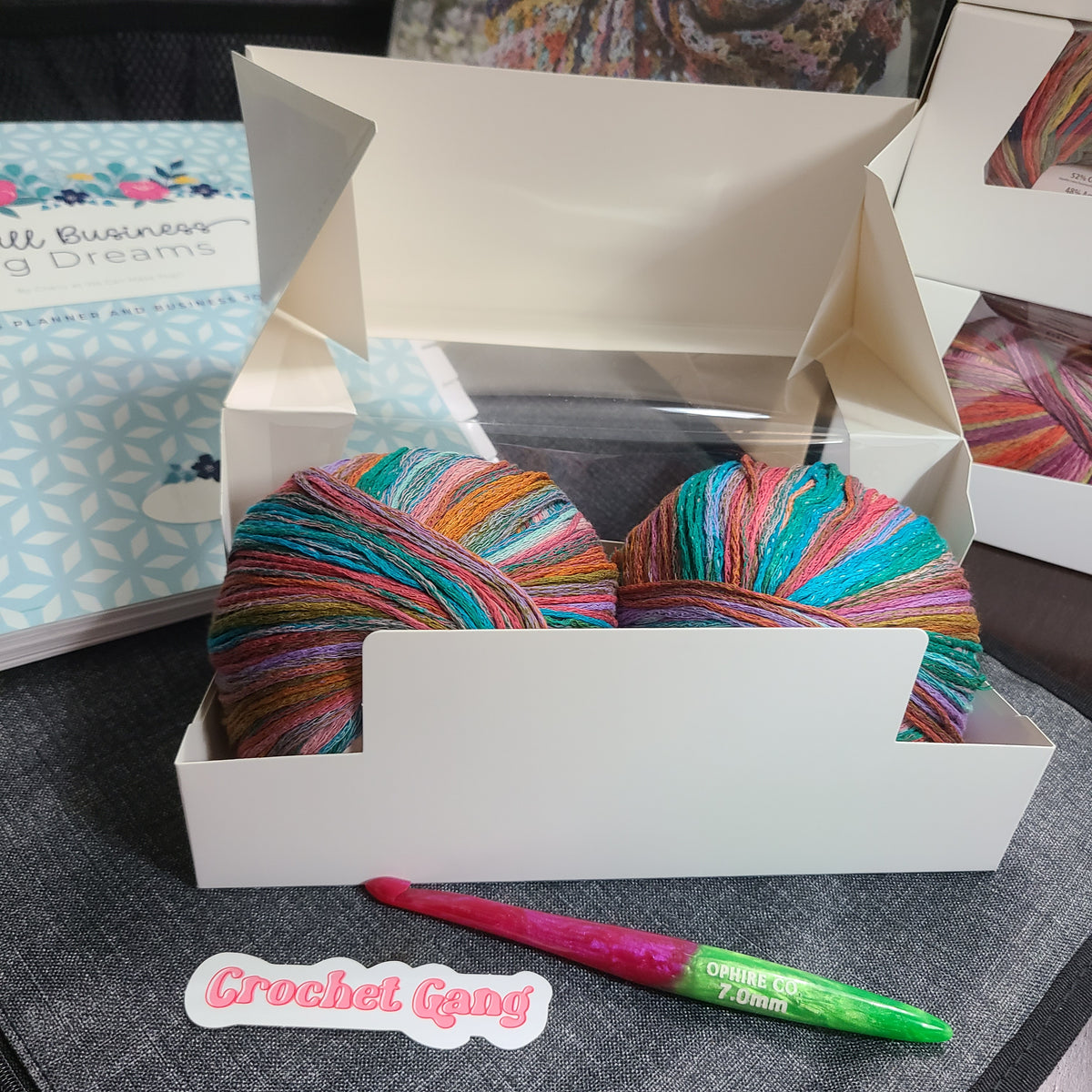 A white cardboard box containing two skeins of multicolored crochet yarn, with a pink and green crochet hook and a 'Crochet Gang' sticker placed in front on a dark gray surface.