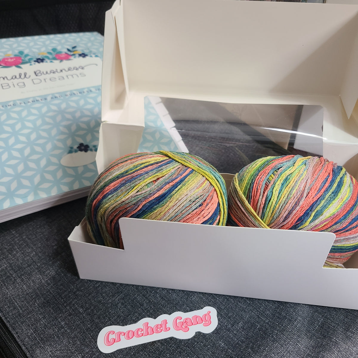 Two balls of variegated crochet yarn in a white cardboard box, with a small 'Crochet Gang' sticker in front and a book in the background.