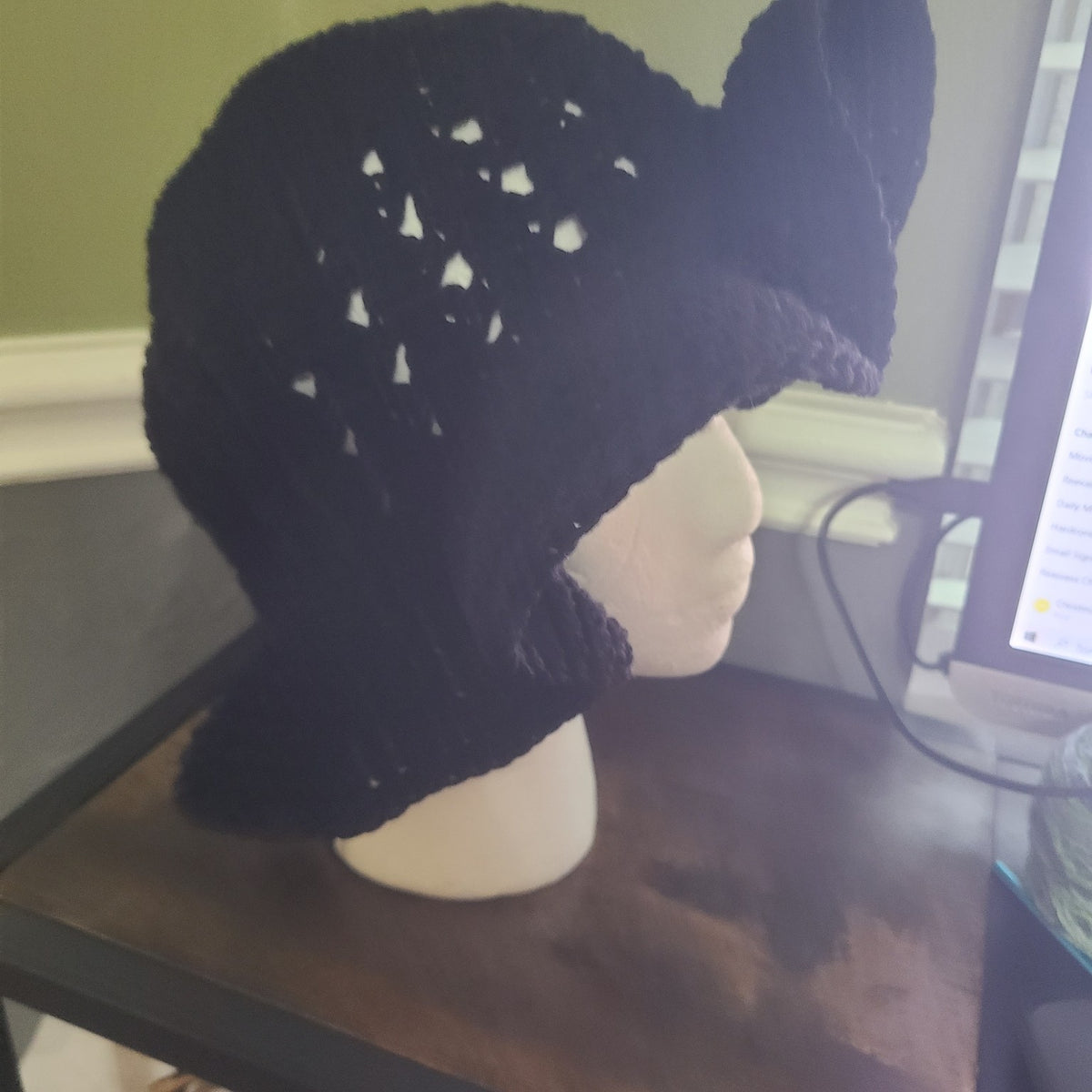 A black knit sun hat with a wide brim and ear flaps, displayed on a white mannequin head on a wooden table in an indoor setting.