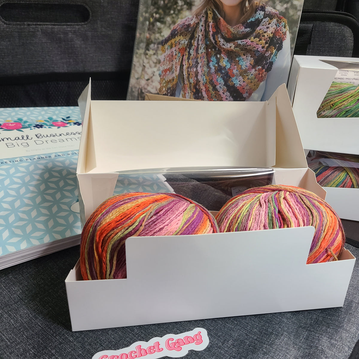 Two skeins of variegated crochet yarn in bright orange, pink, purple, green, and yellow tones, displayed inside an open white cardboard box. A booklet and a printed photo of a crocheted shawl are visible in the background on a dark gray fabric surface.