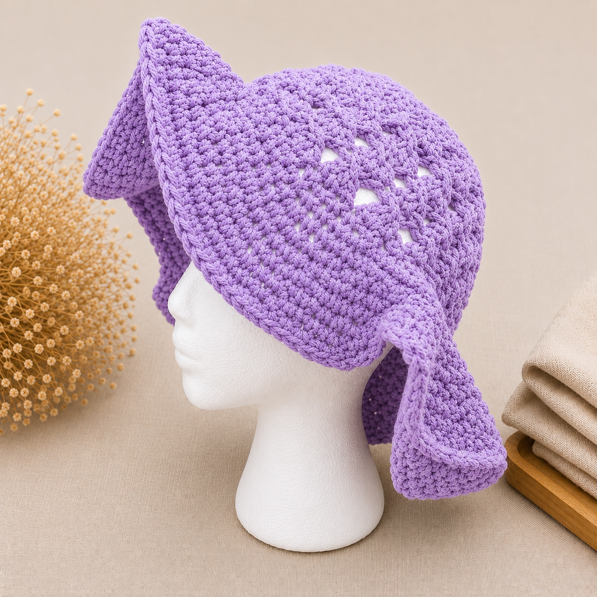 A lavender purple crocheted sun hat with floppy ear flaps, displayed on a white mannequin head. The background is a warm beige with decorative dried flowers on the left and folded fabric on the right.