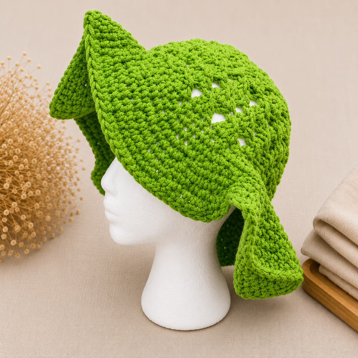 A bright green crocheted hat with floppy ear flaps, displayed on a white mannequin head against a warm beige background with dried flowers and folded fabric props.