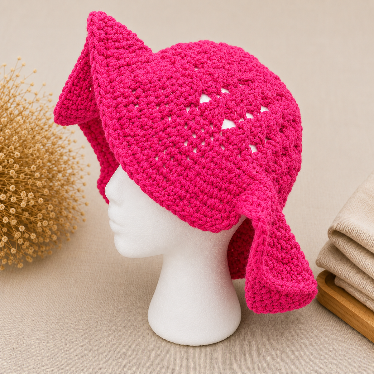 A bright pink crocheted hat with ear flaps and triangular cutout details, displayed on a white mannequin head against a beige background with decorative dried flowers and folded fabric.