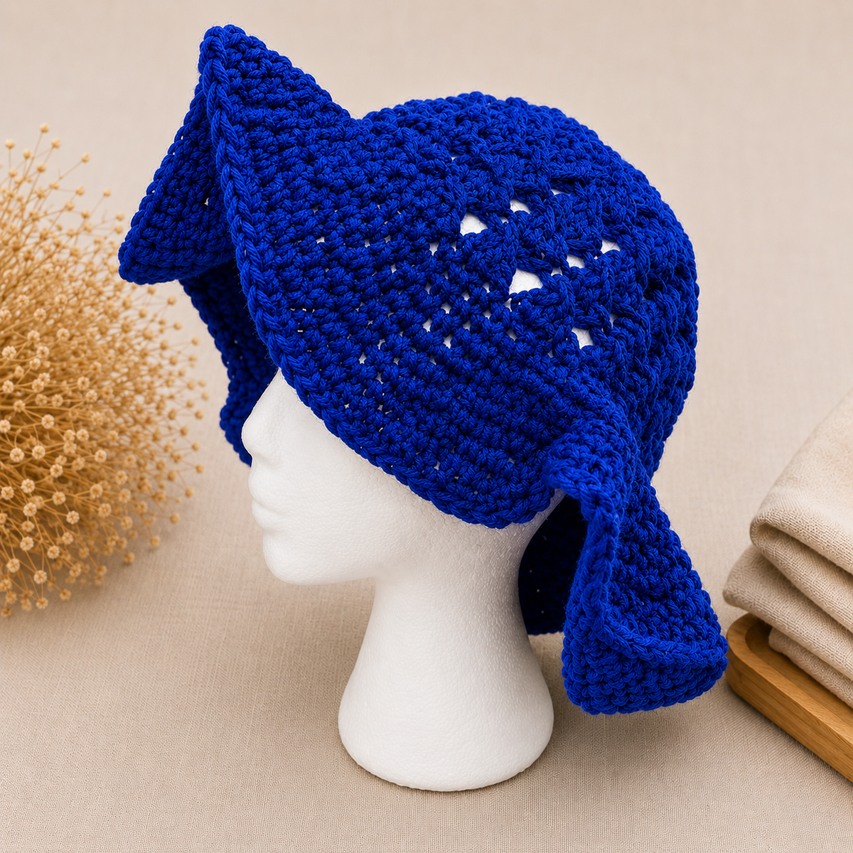 A bright royal blue crocheted hat with ear flaps and a pointed top, displayed on a white mannequin head against a warm beige background with decorative dried flowers and folded fabric.