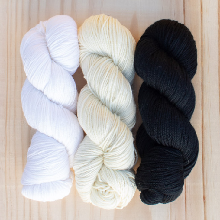 Three skeins of yarn in white, cream, and black on a wooden surface