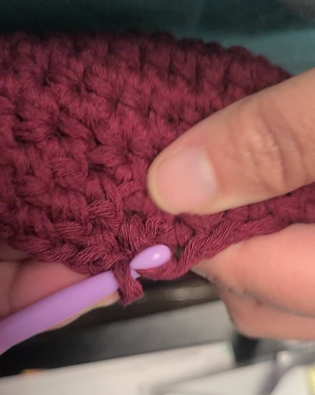 Close-up of a hand holding a maroon crochet hat and purple crochet hook.
