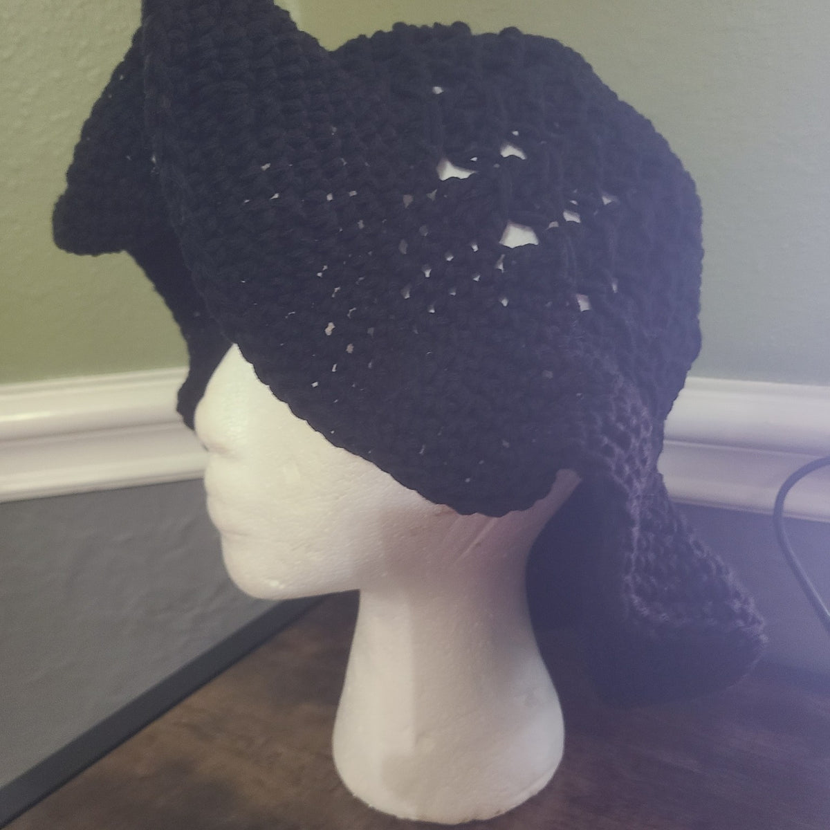 A black crocheted hat with a wide brim and long draped side flaps, displayed on a white mannequin head. The hat features an openwork, textured crochet pattern with small white highlights showing through.