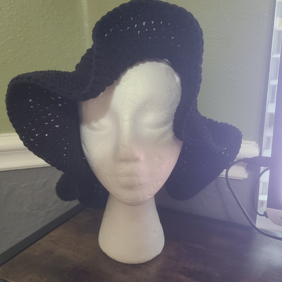 A black crocheted sun hat with a wide brim displayed on a white styrofoam mannequin head, set on a dark wooden table against a light green wall.